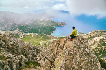 Trourist boy sitting on the rock mountain high above the sea and looking down Corsica Corse France