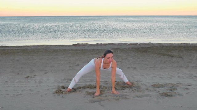 Young woman doing yoga sitting in front splits on the sandy beach at sunrise.