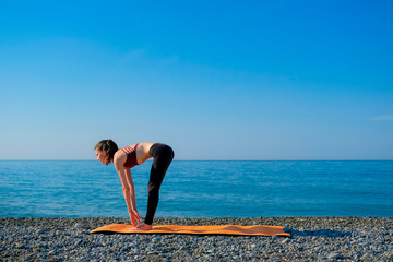 Fototapeta na wymiar Young slim woman in tight sportswear standing on orange yoga mat and practicing outdoors at pebble beach by the sea. Yoga at nature concept