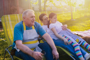 Fototapeta na wymiar Older couple sitting with their granddaughter in backyard and making barbeque. Family time.