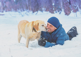 A happy man with a Labrador retriever dog lying in the snow in winter
