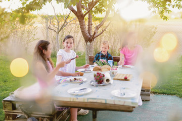 Picture of four kids sitting by the table in nature and eating. Beautiful summer day.