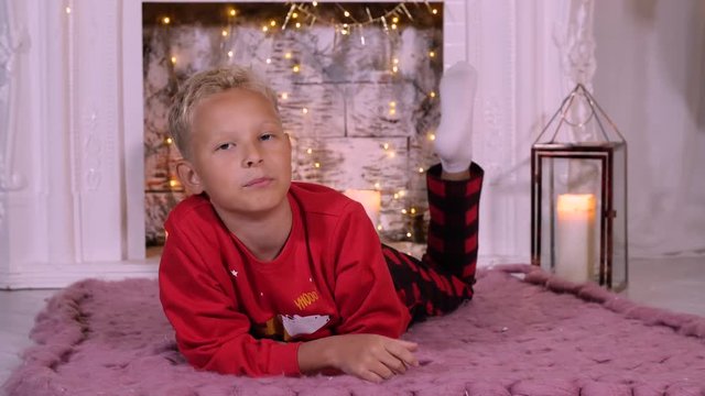 Young blonde boy lye at floor on christmass fireplace background interior. Red pullover. Caucasian white young boy. Xmass lantern