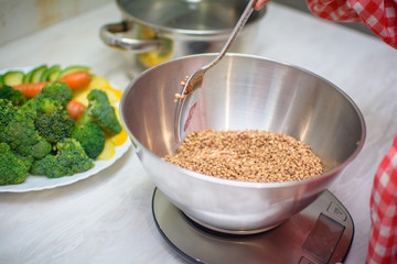 cute baby weighs buckwheat and vegetables on kitchen scales. A little girl is cooking vegetarian food in her kitchen. Healthy eating concept
