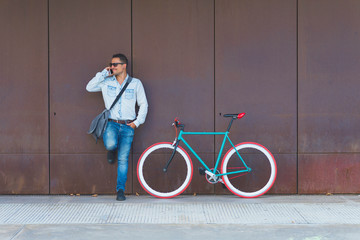 Man talking on smart phone while standing by bicycle against metallic wall at sidewalk in city