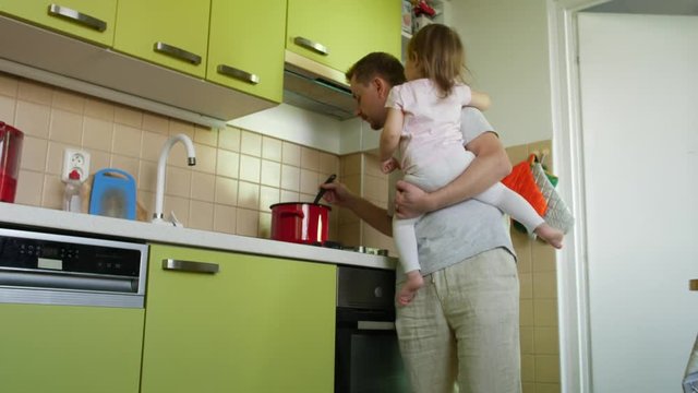 Father's day. Cooking father holding small daughter in arms lauds son for homework in kitchen. Dad making a meal, taking care of small girl and praising elder boy for lessons. 2 children and parent