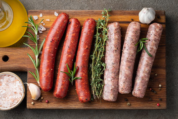 Assorted fresh sausages with thyme, rosemary, olive oil and garlic on a wooden chopping Board on a dark concrete table. Ready to cook sausages for dinner on brown background. Top view with copy space