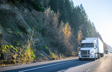 White big rig semi truck with dry van semi trailer driving in front of another semi trucks and cars traffic on wide highway