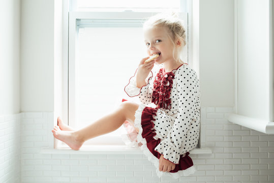 Young girl sitting on a window sill eating a cookie