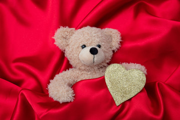 Valentines day. Top view of a teddy bear and golden heart, on red silk background.
