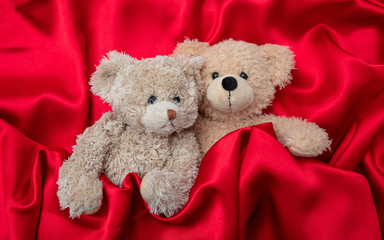 Love, concept, tight hug. Two teddy bears embracing as a couple in bed, red satin background