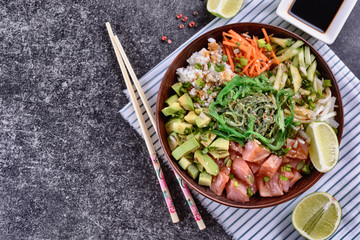 Homemade healthy Poké bowl with rice, salmon, avocado, white onion, scallion greens, sesame oil, soy and sesame sauce, seaweed (wakame) and other ingredients on a dark backgound are ready to eat