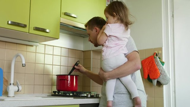 Father's day. Cooking father with small daughter in hands yelling at son for homework in kitchen. Dad making a meal, taking care of small girl and berates for lessons elder boy. 2 children and parent
