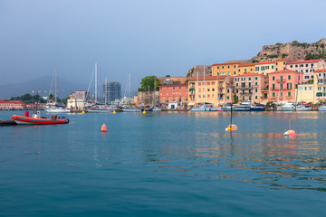 Beautiful view of the port of the city Portoferraio. Bright red boat in the foreground and colorful houses on the waterfront in the background under a dark rainy sky.