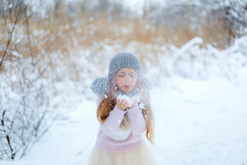 Pretty little girl wearing grey knitted hat and pink sweater walking in snowy park.  Cute child blowing snowflakes in winter forest. Family vacation with kid on christmas holidays. Blurred background