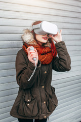 Pretty hipster young girl plays virtual reality mobile game app on smartphone attached to modern VR glasses outdoor in cold winter day