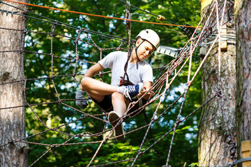 Naklejka na ściany i meble Child in forest adventure park. Kid in white helmet and white t shirt climbs on high rope trail. Agility skills and climbing outdoor amusement center for children. young boy plays outdoors