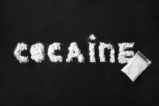 Word Cocaine written with white powder and full plastic bag on black background, top view