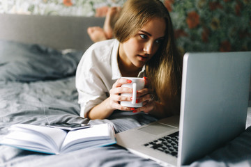 Attractive smiling european woman using laptop and drinking coffee while lying on her bed. Young woman drinking coffee at home, start, concept in the morning, early in the morning. Stylish Toned