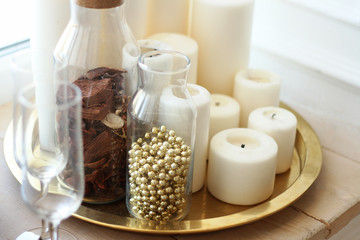 composition from white candles, transparent small bottles and crystal glasses on a gold tray stand on a window sill