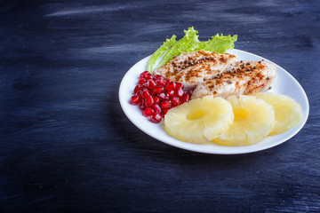 Fried chicken fillets with lettuce, pineapple and pomegranate seeds on black wooden background.