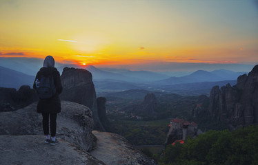 Fototapeta premium Happy woman stay on the peak of the mountain cliff edge under sunset light sky enjoying the success, freedom and bright future. Monastery Meteora Kalambaka in Greece on background.
