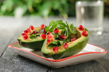 A glass of clean water with avocado halves filled with pomegranate seeds and parsley on a wooden table. Vegetarian cuisine for weight loss.