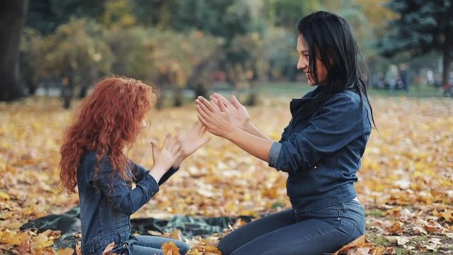 Little redhead girl with her mommy spend time in autumn park. They play and clap hands