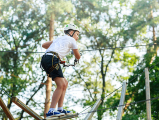 Cute boy enjoying activity in climbing adventure park at sunny summer day. Kid climbing in rope playground structure. Safe climbing with helmet insurance. Child in forest adventure park, extreme sport