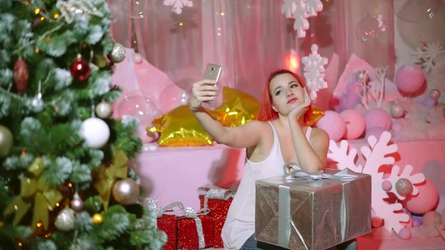 Beautiful young girl with red hair taking selfie on smartphone with a christmas present.