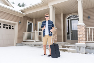 Excited young traveler with his suitcase leaving his home for a winter vacation. He is wearing shorts and a t shirt while standing in the snow.