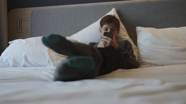 The boy is lying on the bed with a smartphone in his hands. Communication and games on the smartphone