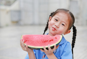 Happy little Asian child girl in school uniform enjoy eating watermelon outdoors.