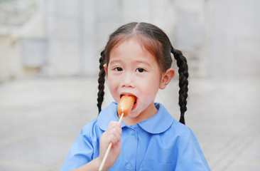 Happy little Asian child girl in school uniform enjoy eating sausage outdoors.
