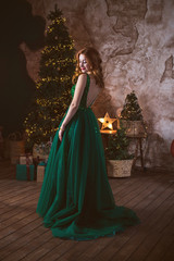Fototapeta premium Winter holidays, celebration and people concept - young sexy woman in elegant green evening dress over christmas interior background