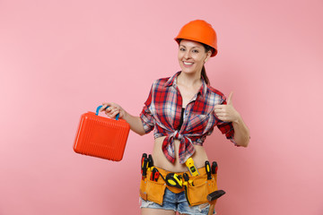 Strong young handyman woman in orange helmet, plaid shirt, denim shorts, kit tools belt full of instruments, toolbox isolated on pink background. Female in male work. Renovation occupation concept