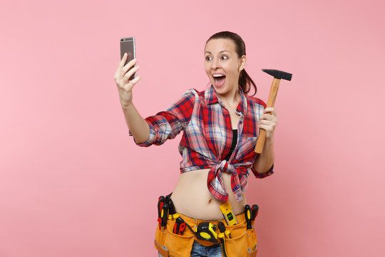 Overjoyed fun handyman woman in plaid shirt, denim shorts, kit tools belt full of instruments, hammer doing selfie on mobile phone isolated on pink background. Female in male work. Renovation concept.