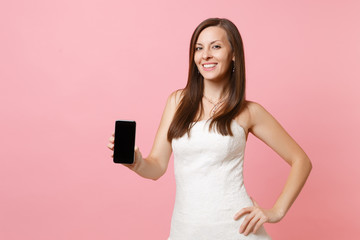 Portrait of smiling bride woman in wedding dress holding mobile phone with blank black empty screen isolated on pastel pink background. Wedding to do list. Organization of celebration. Copy space.