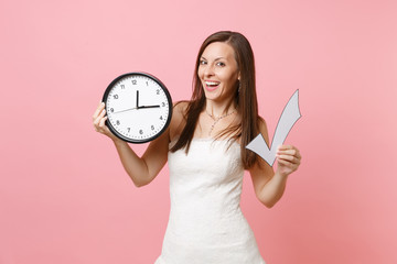 Smiling bride woman in lace wedding dress holding check mark and round alarm clock isolated on pastel pink background. Time is running out. Wedding to do list. Organization of celebration. Copy space.
