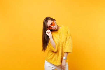 Pretty upset young woman in fur sweater and white pants holding heart orange glasses, blowing lips isolated on bright yellow background. People sincere emotions, lifestyle concept. Advertising area.