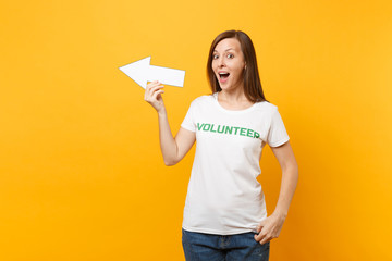 Portrait of happy woman in white t-shirt written inscription green title volunteer hold arrow pointing aside isolated on yellow background. Voluntary free assistance help, charity grace work concept.