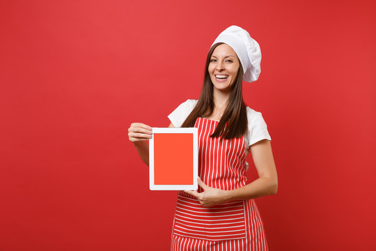 Housewife female chef cook or baker in striped apron white t-shirt toque chefs hat isolated on red wall background. Woman hold tablet pc blank screen for promotional content Mock up copy space concept