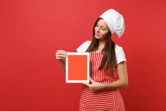 Housewife female chef cook or baker in striped apron white t-shirt toque chefs hat isolated on red wall background. Woman hold tablet pc blank screen for promotional content Mock up copy space concept