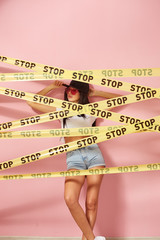 Young dark-haired girl in pink sunglasses and cap dressed in jeans shorts and white top stands behind the yellow scotch tapes with inscriptions of word 