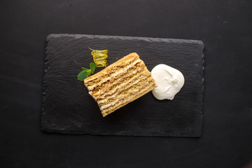 Honey cake piece with cream on stone plate on black background
