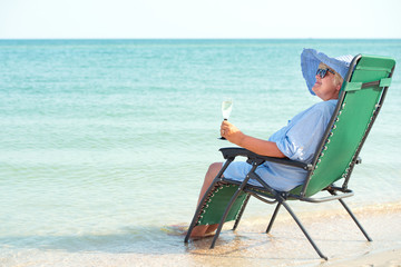 An elderly woman sits on the beach on a chaise longue and drinking wine.	