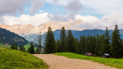 Gravel path on a meadow near the huts and forest in a valley with high sharp mountains in the background in Dolomites, Italy