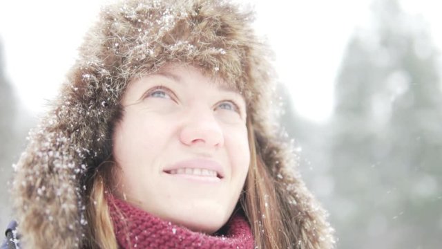 A young woman in a fur hat with earflaps dreamily looks at the sky. On the street, winter, snow falls. She dreams of a miracle. Close-up on face and headgear