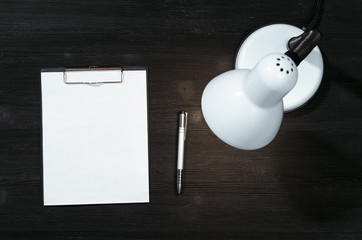 Blank paper page document with copy space and lamp on the black office table background. To do...