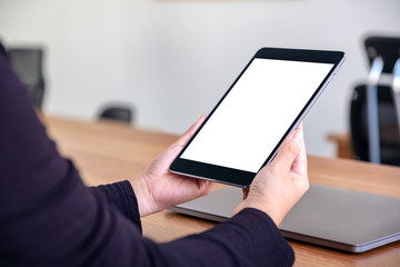 Mockup image of hands holding and using black tablet pc with blank white desktop screen with notebook on wooden table in office